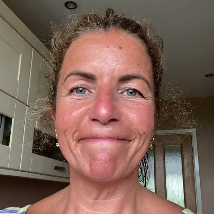 A woman with curly hair and a friendly expression is standing in a kitchen, looking directly at the camera. Her expression and welcoming, suggesting a positive testimonial.