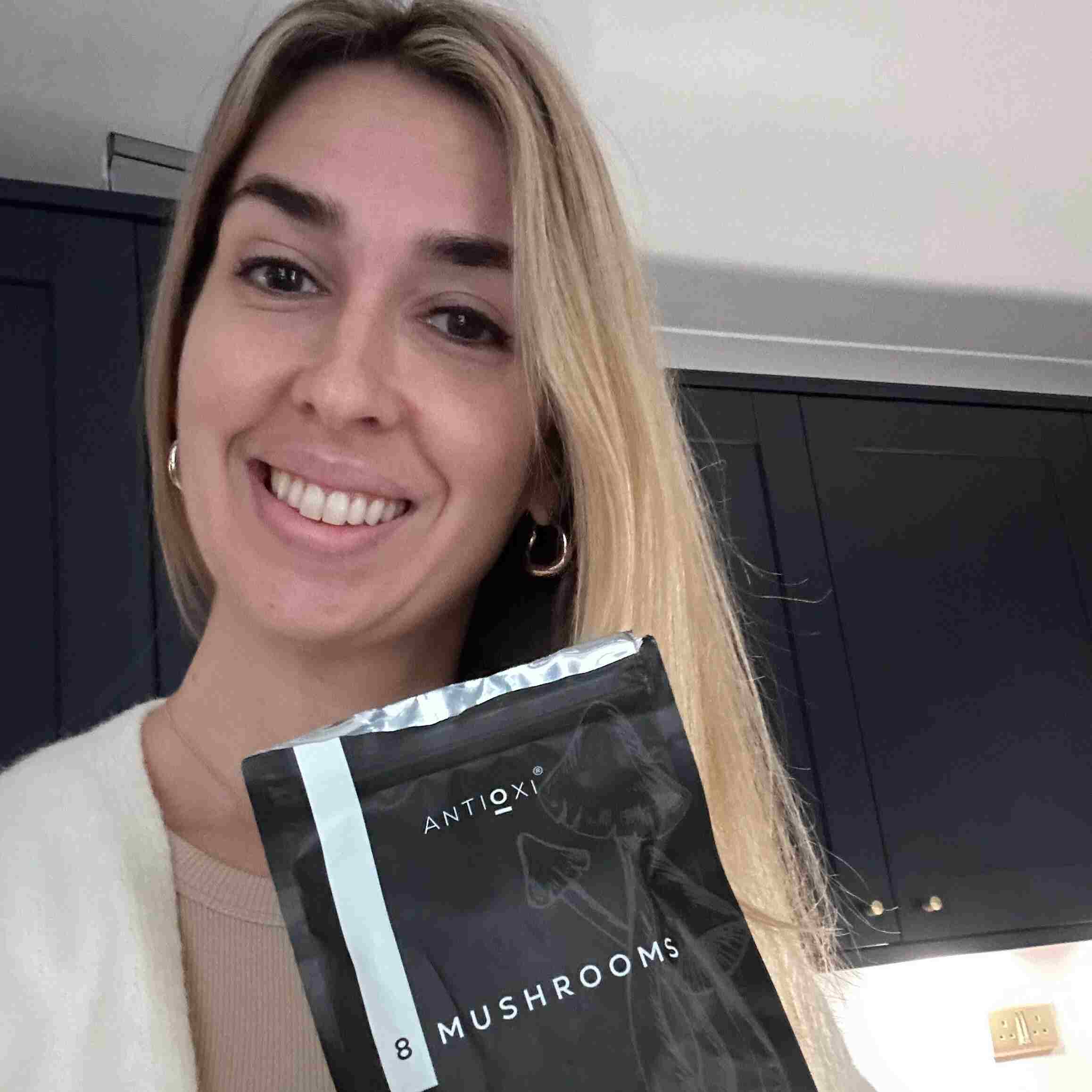 A woman with blonde hair and a bright smile is standing in a kitchen, looking directly at the camera and holding a pouch of Antioxi 8 Mushrooms Blend mushroom supplement. Her expression is friendly and welcoming, suggesting a positive testimonial.