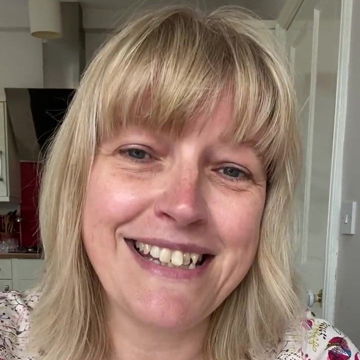 A woman with blonde hair and a bright smile is sitting in a kitchen, looking directly at the camera. Her expression is friendly and welcoming, suggesting a positive testimonial.