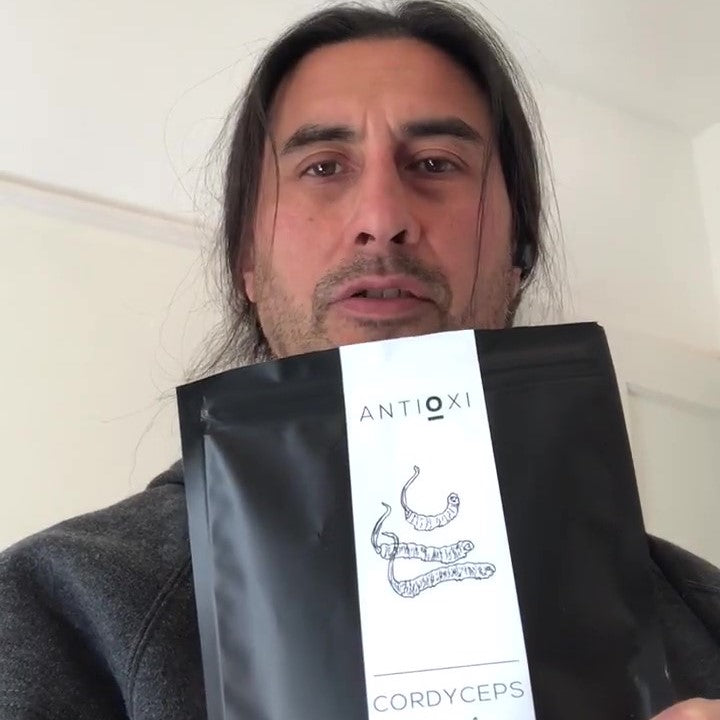 A man with a friendly expression is standing indoors, giving a review of Antioxi Cordyceps mushroom supplements.