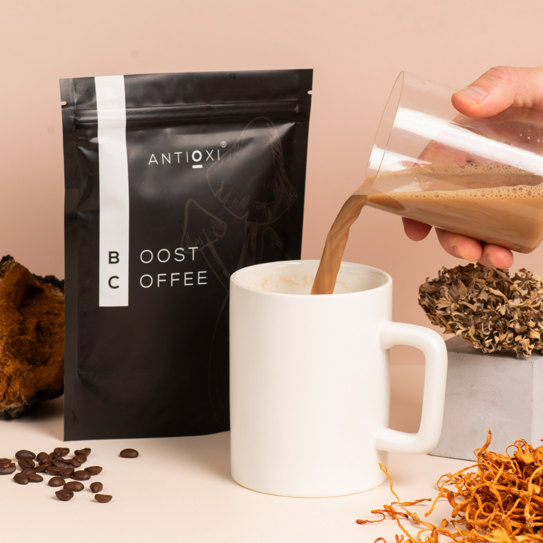 A pouch of Antioxi Boost Mushroom Coffee is displayed alongside a white mug, with coffee being poured into the mug. Surrounding the setup are scattered coffee beans and dried Cordyceps, Maitake and Chaga mushrooms.