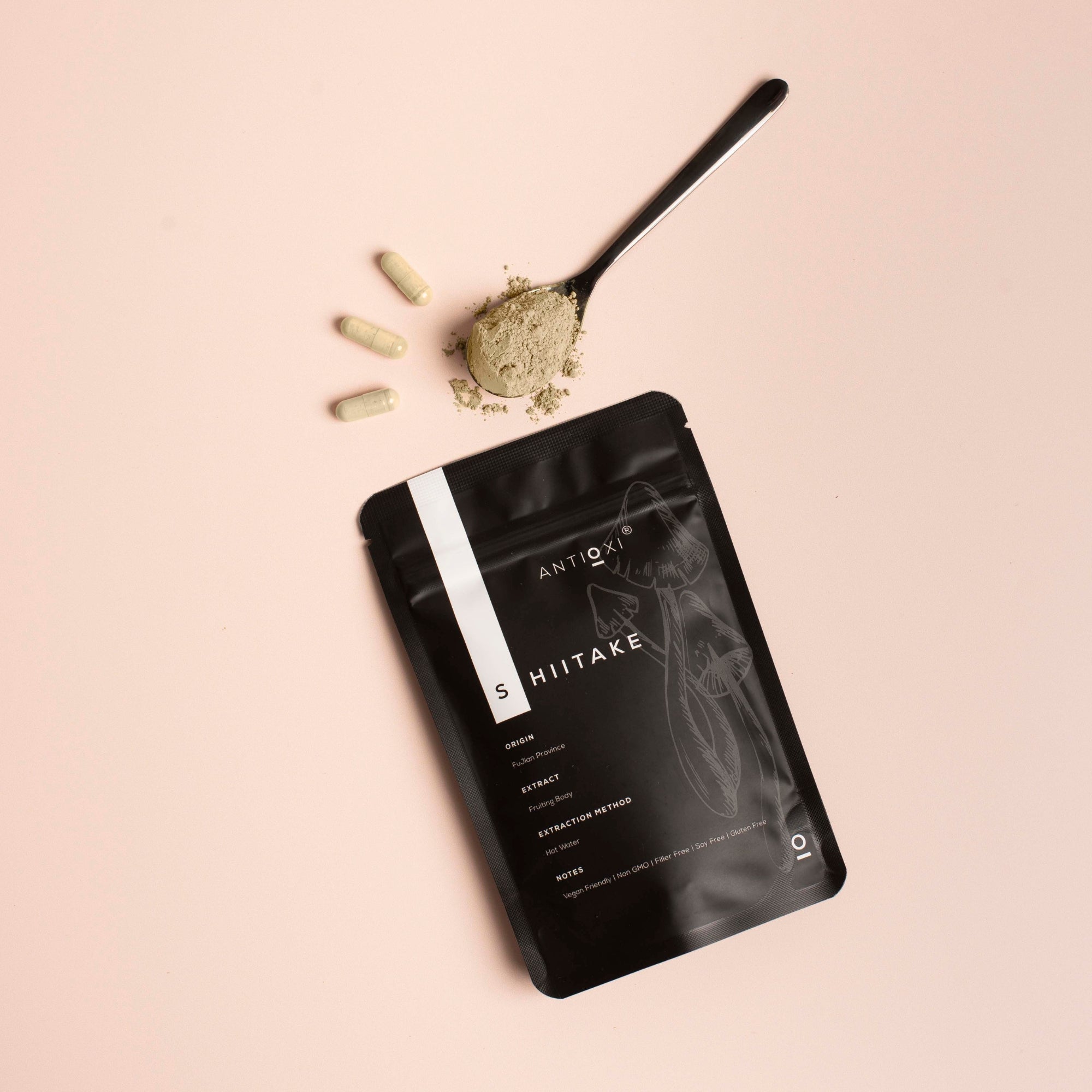 A pouch of Antioxi Shiitake mushroom supplements is placed on a light pink surface, accompanied by three capsules and a spoonful of Shiitake powder.