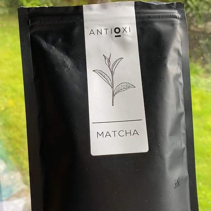 Antioxi Matcha package displayed against a green, natural background, showcasing the product's minimalist design with a white label displaying a simple illustration of a tea leaf.