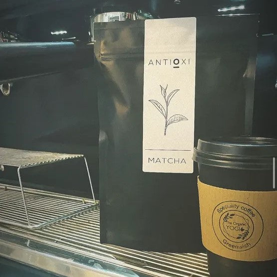 Antioxi Matcha powder pouch next to a takeaway coffee cup on a café counter.