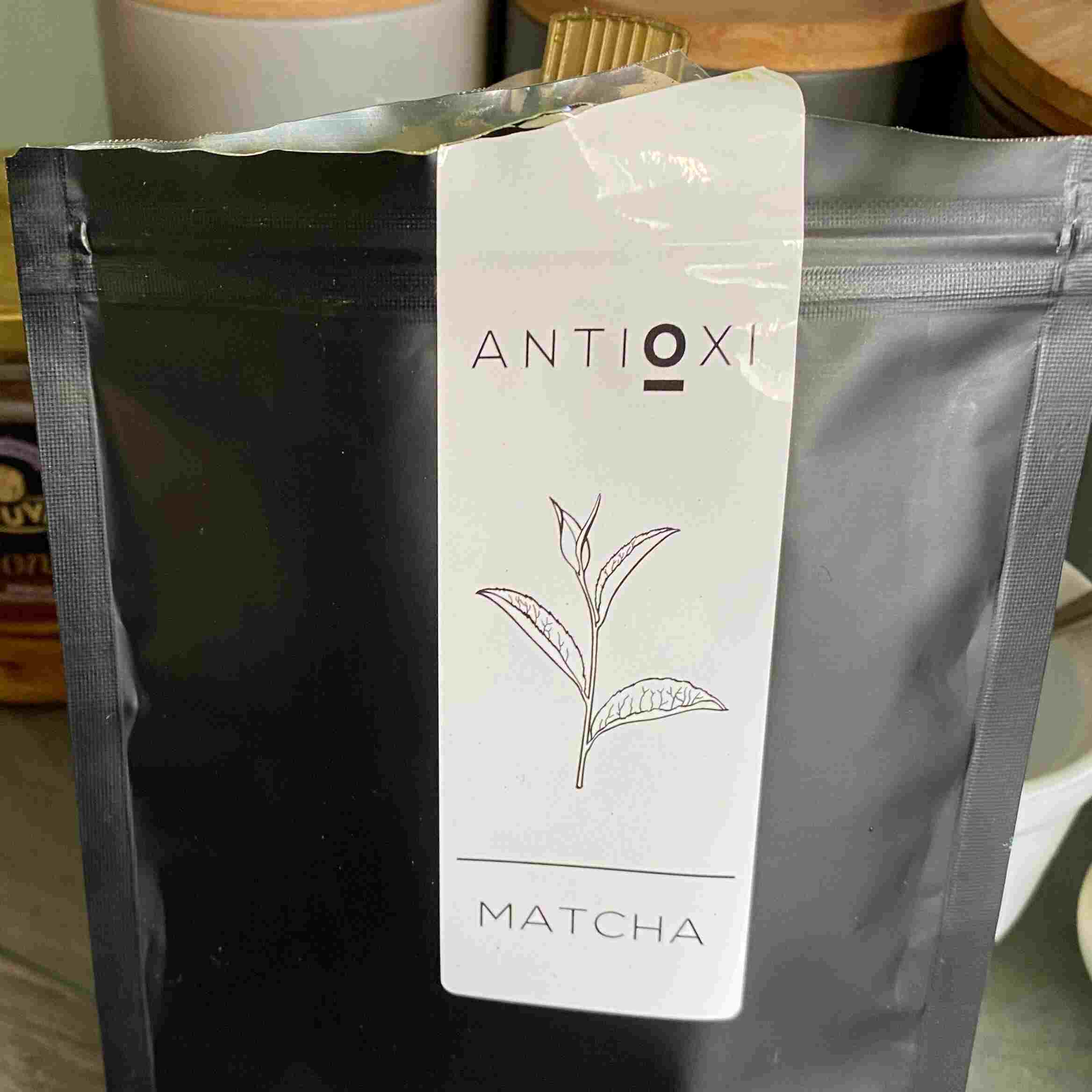 A close-up of a pouch of Antioxi Matcha powder placed on a kitchen counter, emphasizing the product's sleek black packaging and clear labeling with an illustration of a tea leaf.