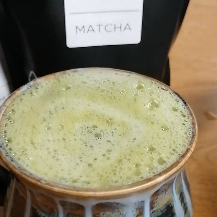 A close-up of a frothy cup of coffee made with Antioxi Matcha powder, featuring a partially visible Antioxi Matcha powder pouch in the background.