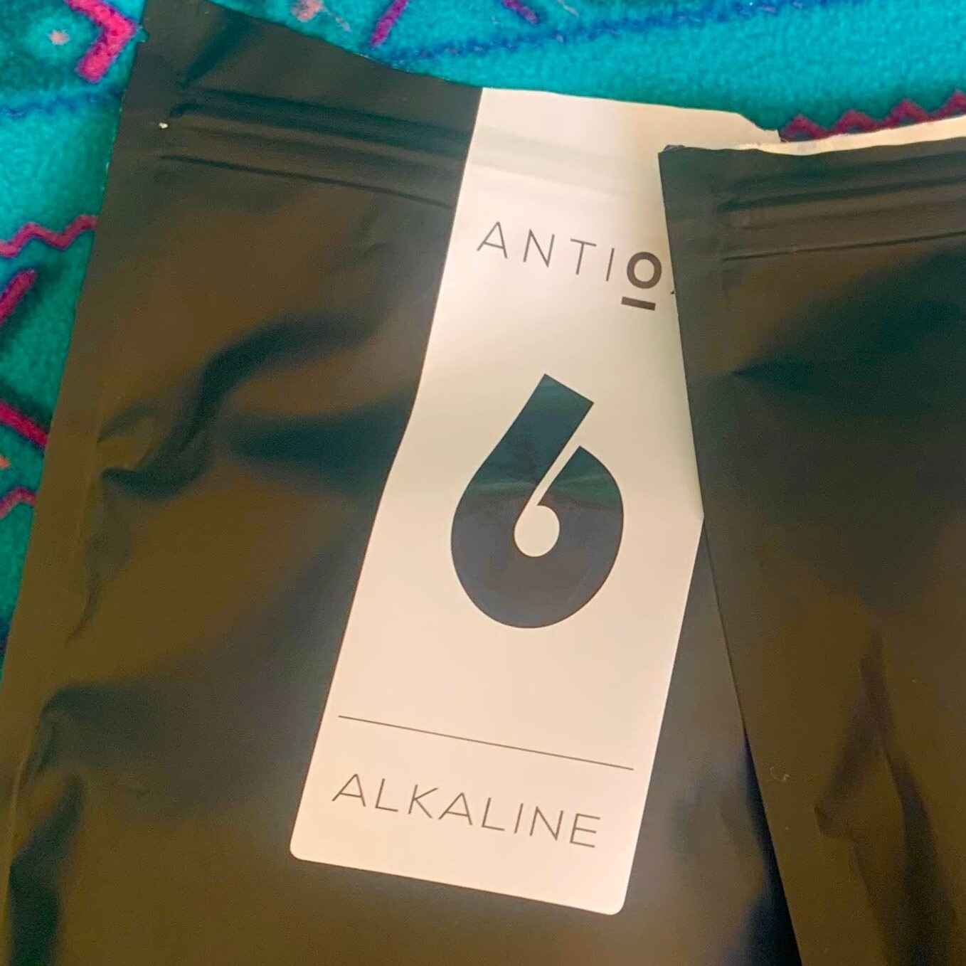 A close-up of a pouch of Antioxi Alkaline Greens blend placed on a soft, blue surface, with the side of another pouch partially visible next to the Alkaline Greens Blend.