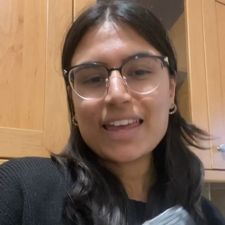 A woman with black hair and a bright smile is standing in a kitchen, looking directly at the camera. Her expression is friendly and welcoming, suggesting a positive testimonial.