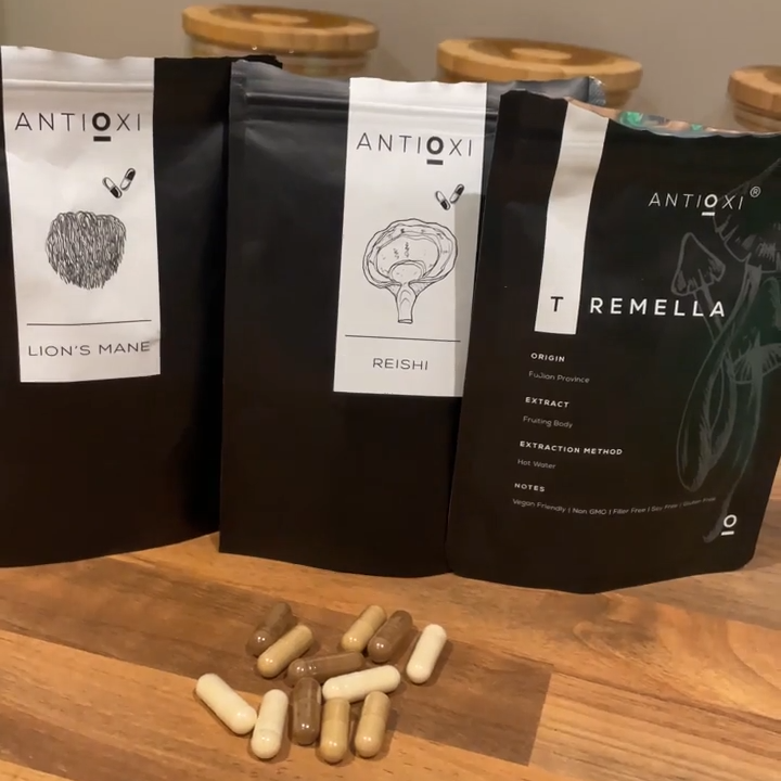 Pouches of Antioxi Lion's Mane, Reishi and Tremella mushroom supplements placed on a wooden surface, with a mix of capsules placed in front of the pouches. The capsules each contain different coloured powders, namely white for Tremella, light yellow for Lion's Mane and brown for Reishi.