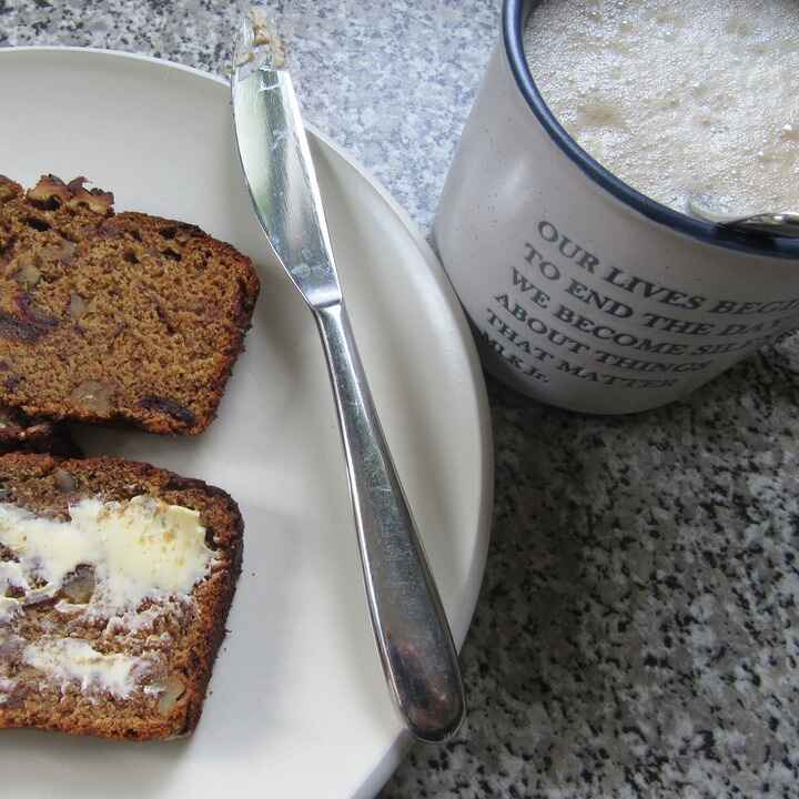 Slices of nut bread with butter on a plate next to a mug of foamy coffee made with Antioxi Focus mushroom coffee powder.