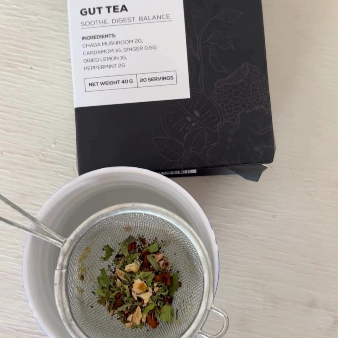 Antioxi Gut Mushroom Tea box placed on a white surface, next to a cup with tea strainer filled with loose leaf ingredients placed on top of the cup.