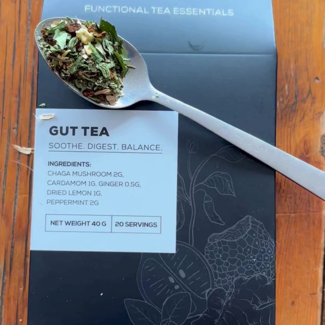 A close-up of Antioxi Gut Mushroom Tea tea-box laying down on a wooden surface with a spoonful of loose leaf ingredients placed on top of the box.